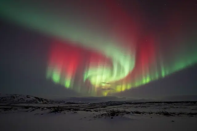 Vibrant red and green aurora arcs across a dark starry sky above a snow-covered northern tundra landscape.