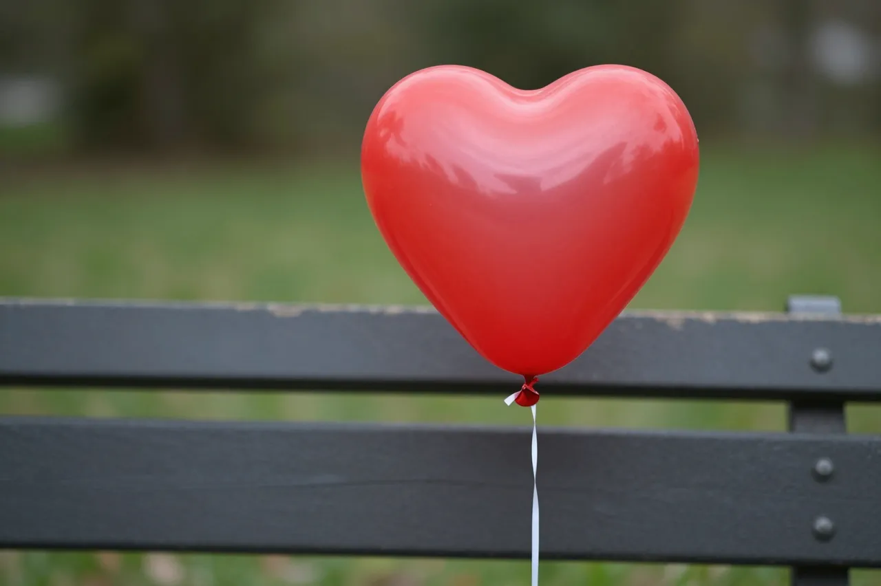 A shiny red heart-shaped balloon rests in front of a dark wooden park bench with blurred park grass in the background.