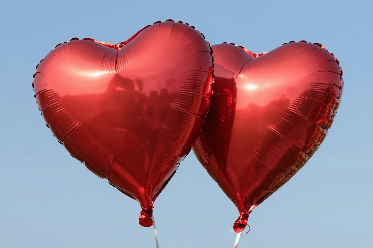 Two shiny metallic red heart-shaped balloons float against a clear light blue sky