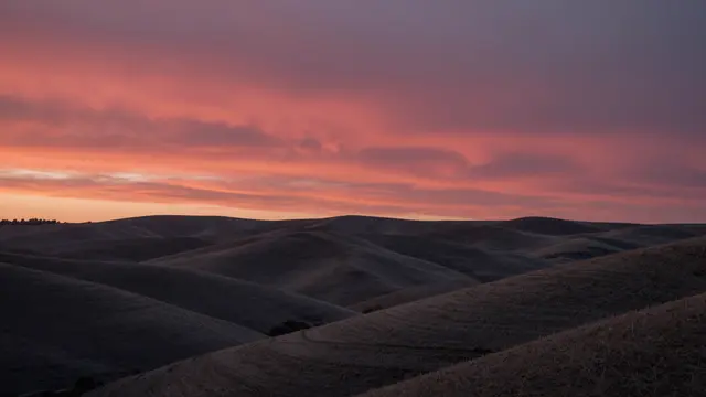 Warm pink and red dawn sky stretches above silhouetted dark rolling grassy hills