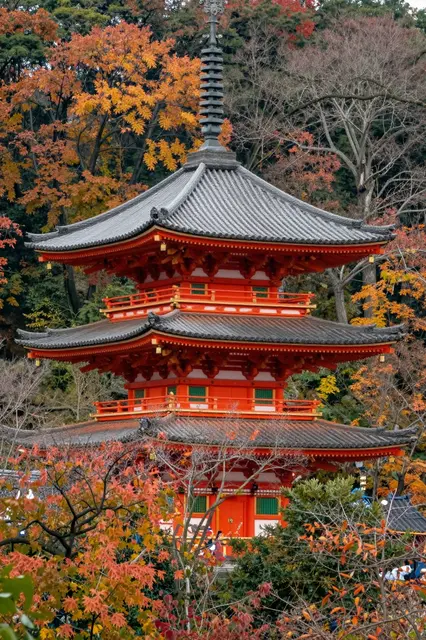 A three-tiered red Japanese pagoda stands framed by vibrant orange and red autumn trees on a forested hillside