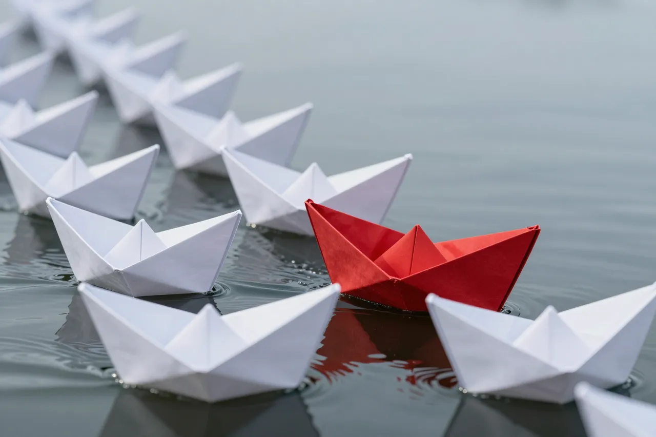 A single red origami paper boat sails at the front of a line of white paper boats on calm still water