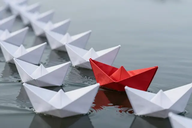 A single red origami paper boat sails at the front of a line of white paper boats on calm still water
