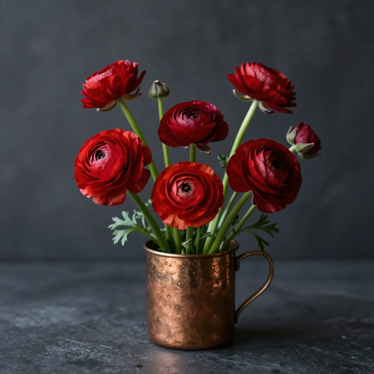 Vibrant red ranunculus blooms sit in a hammered copper mug against a dark charcoal still life backdrop