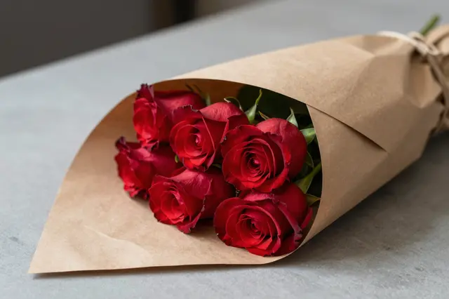 A bouquet of fresh red roses wrapped in plain kraft paper rests on a smooth gray table surface