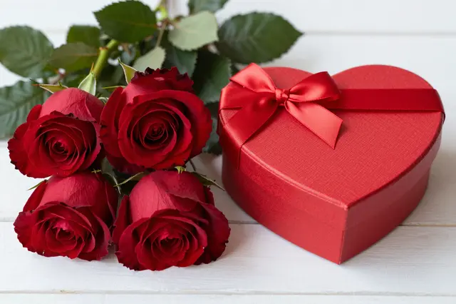 Deep red roses sit next to a satin-bowed heart-shaped gift box on a white wooden surface.