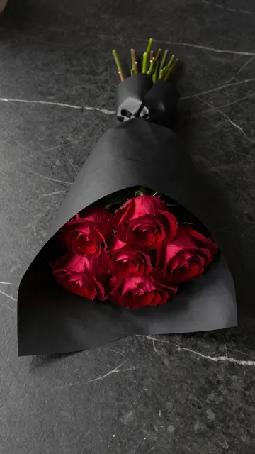 A bouquet of deep red roses wrapped in matte black paper rests on dark marble under moody lighting.