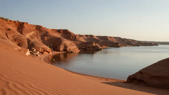 Calm glassy water sits below eroded ochre sand slopes along a quiet red sand cove under a hazy sky
