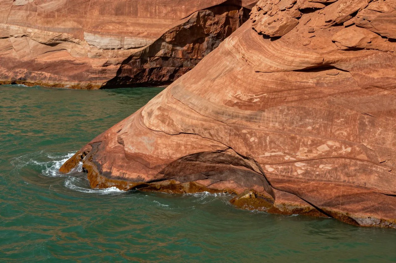 Layered red sandstone cliffs meet rippling green water, with small waves lapping at the rock base.