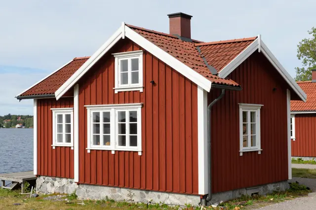 Traditional red wooden cottage with white trim sits beside a calm lake under an overcast sky