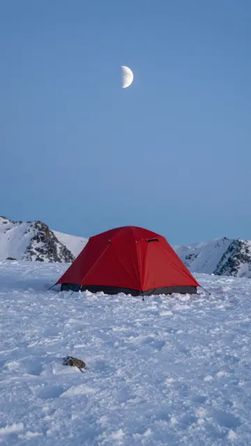 A red camping tent sits on a snow-covered mountain ridge below a crescent moon at blue hour
