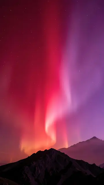 Shimmering red and violet aurora folds glow above dark silhouetted mountain ridges under a starry sky