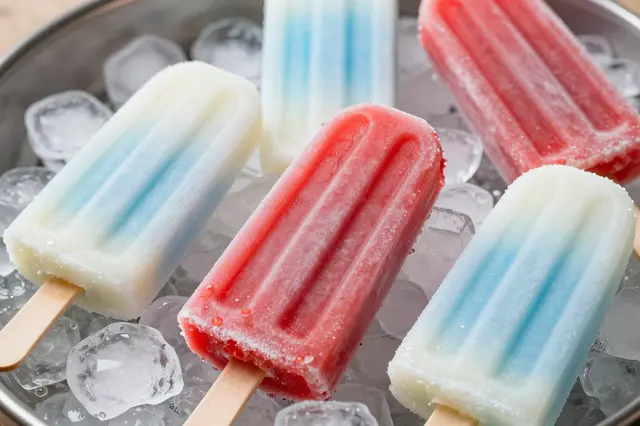 Frosted red, white, and blue popsicles rest on clear ice cubes in a metal tray