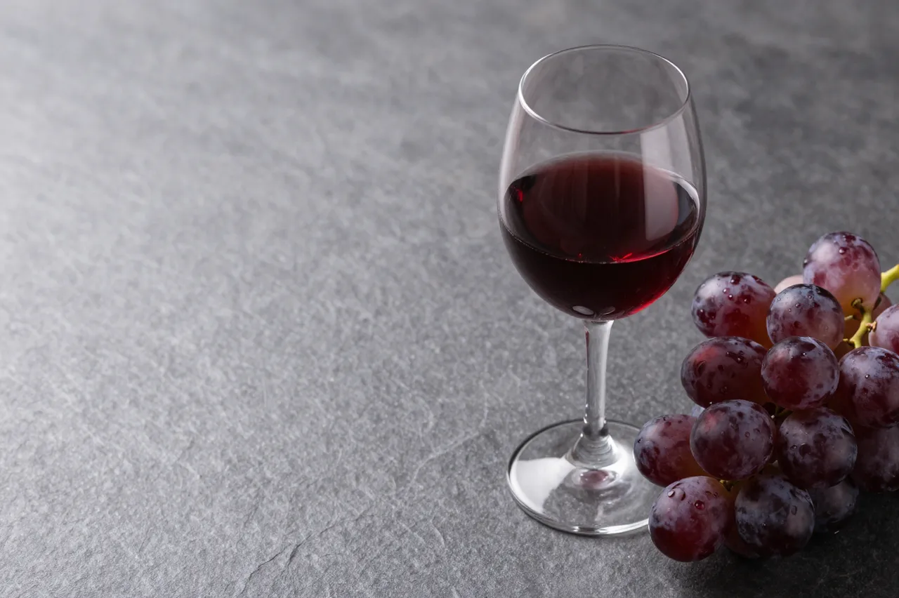 A glass of red wine and a bunch of purple grapes rest on the right of a dark gray stone table with empty left copy space