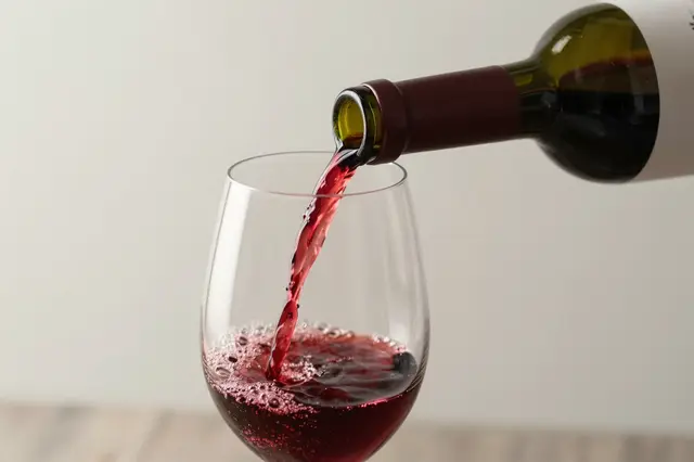 Red wine pours from an unlabeled bottle into a clear stemmed wine glass.