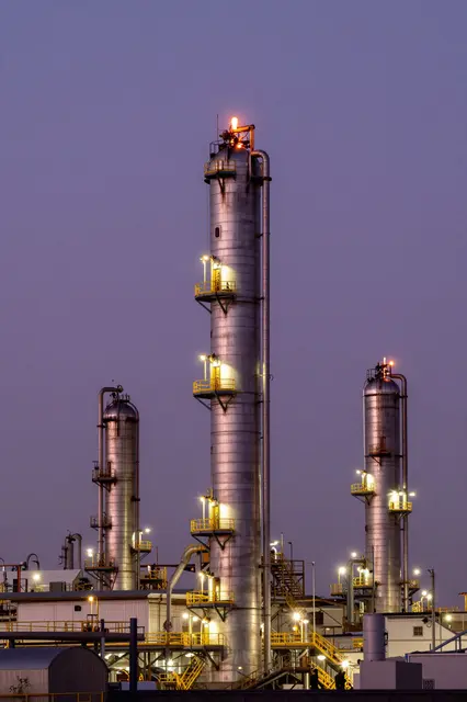 Illuminated refinery flare stacks and process towers stand against a soft violet twilight sky