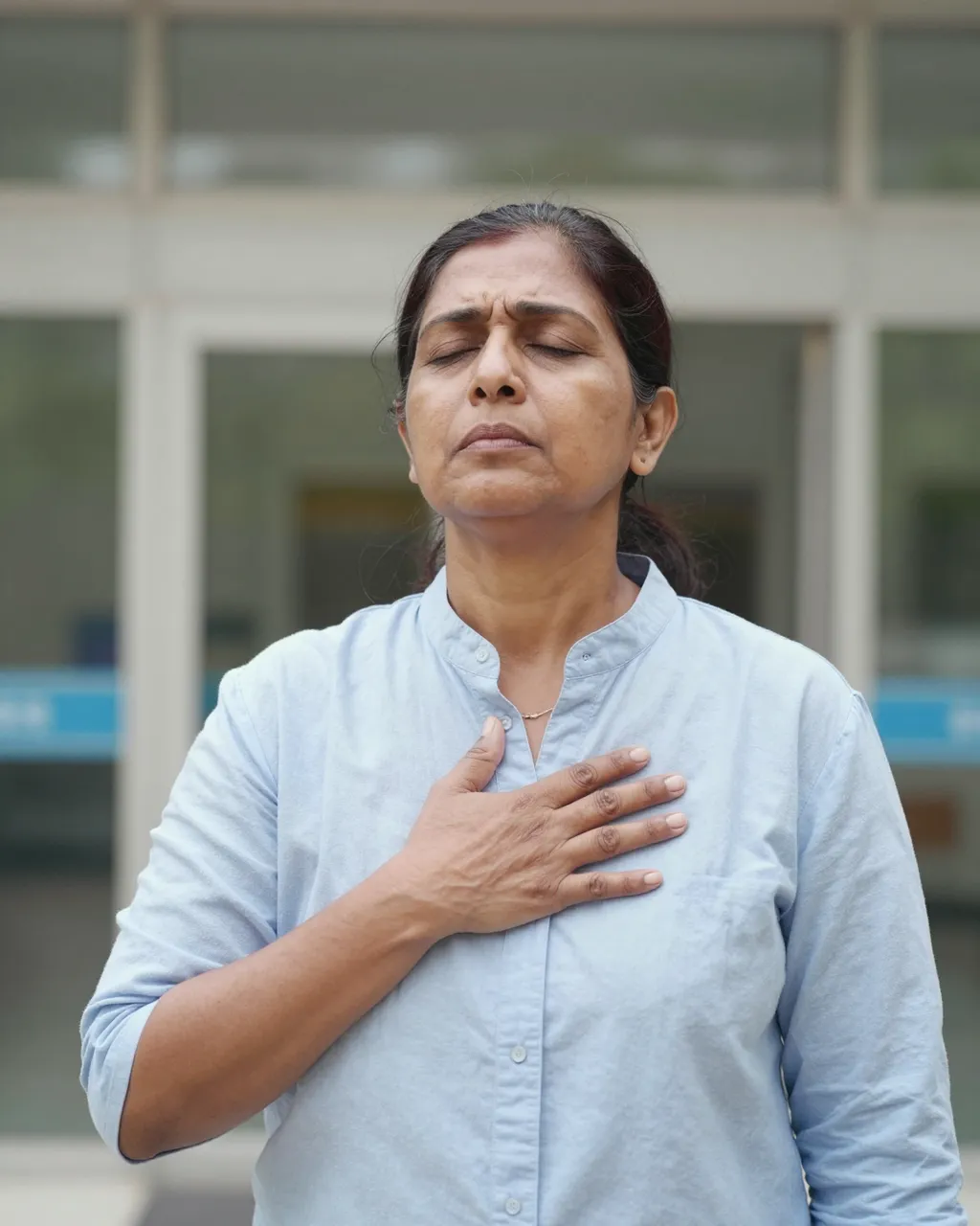 Middle aged South Asian woman stands with eyes closed, hand on her chest outside a hospital building