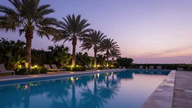 Long resort swimming pool reflects palm trees and soft violet twilight sky at blue hour, lined with glowing garden lights