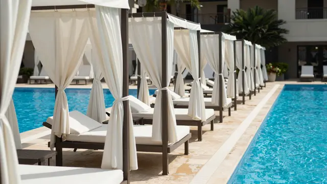 Row of white curtained pool cabanas line the edge of a bright blue resort swimming pool on a sunny day