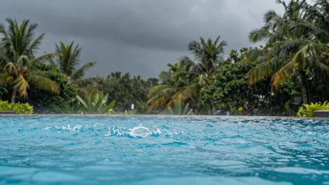 Raindrops splash on a turquoise resort pool under a dark stormy sky with palm trees in the background