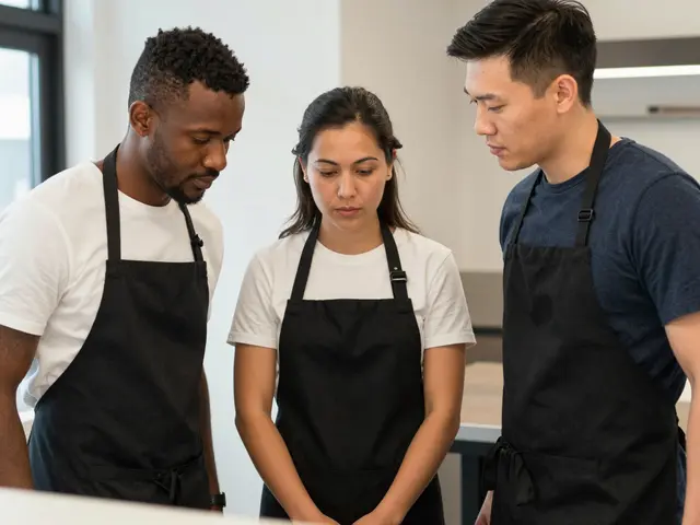 Three diverse restaurant staff in black aprons stand together looking down at a shared off-frame point in a bright kitchen