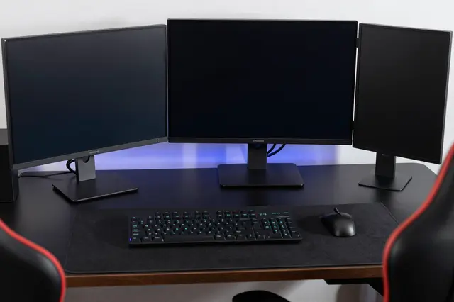 Dark unlit gaming desk with three black monitors, RGB keyboard, mouse, and subtle blue accent lighting