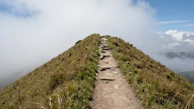 A narrow rocky hiking trail climbs a grassy ridge above cloud-filled valleys under a patch of blue sky
