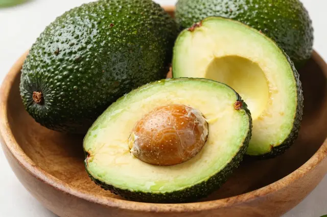 Whole and halved ripe hass avocados rest in a shallow wooden bowl on a light surface