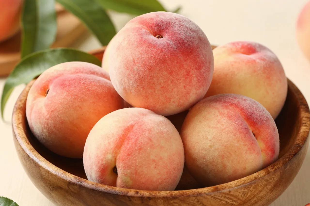 Fresh ripe fuzzy peaches with green leaves piled inside a textured wooden bowl