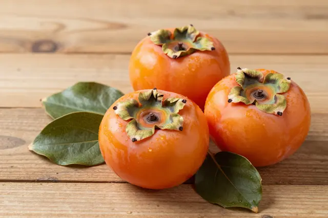 Three glossy ripe persimmons with fresh green leaves rest on a light wooden table surface.