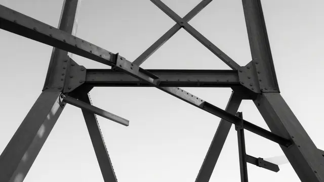 Low angle black and white view of riveted metal bridge girders against a pale overcast sky