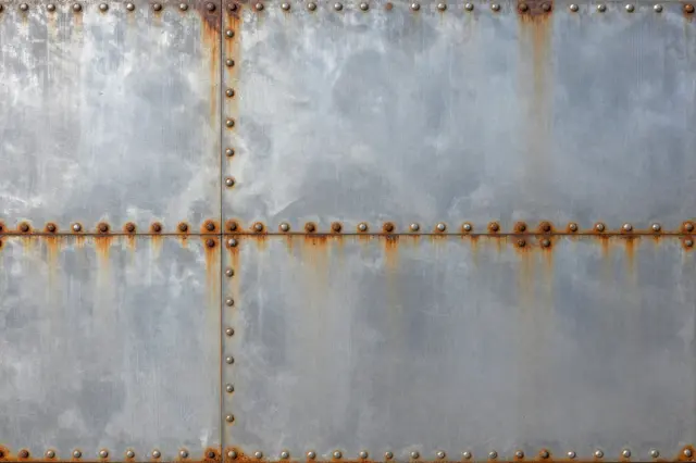 Full frame close-up of a weathered riveted steel panel with rust bleeding around fasteners under flat lighting