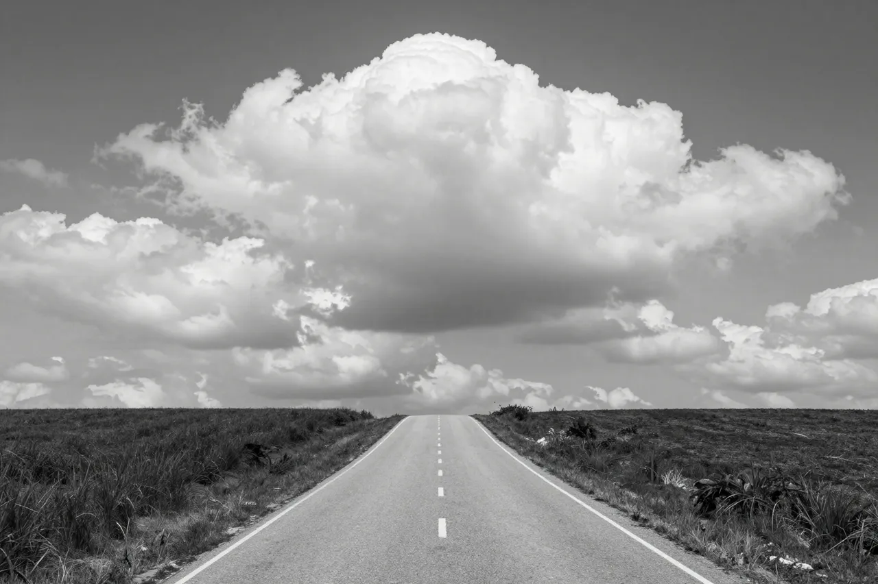 Black and white view of a straight paved country road leading over a hill beneath massive cumulus clouds