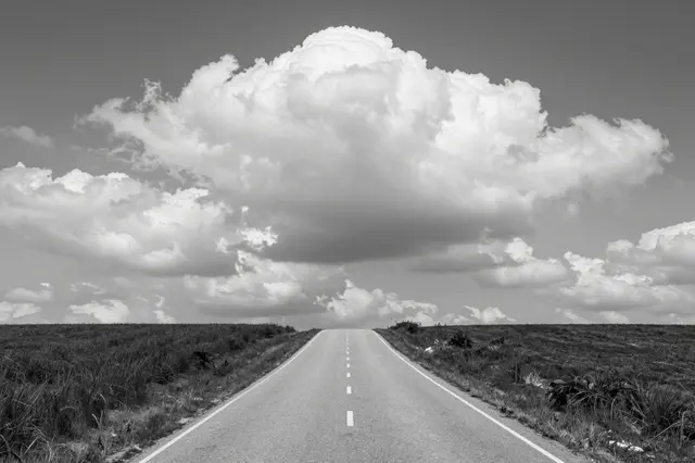 Black and white view of a straight paved country road leading over a hill beneath massive cumulus clouds