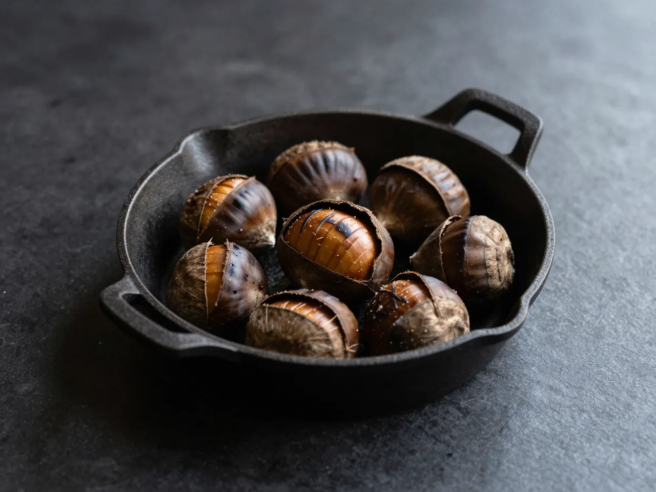 Cracked roasted chestnuts sit in a small cast iron pan on a dark matte tabletop.