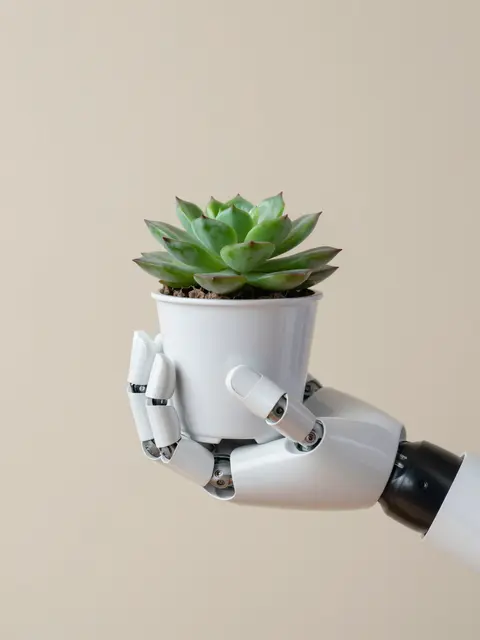 A white robotic prosthetic hand holds a small potted succulent against a soft beige background
