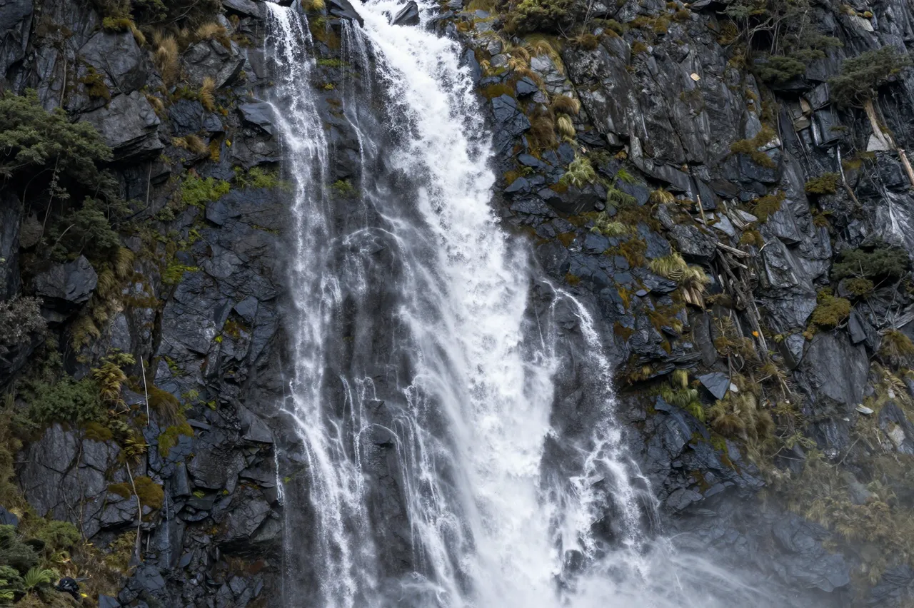 Tumbling waterfalls cascade down a steep dark rocky cliff dotted with moss and small alpine vegetation