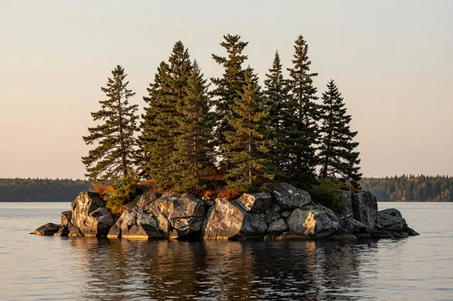 A rocky pine-covered island sits in a calm lake under warm sunset light, with distant forested shores.