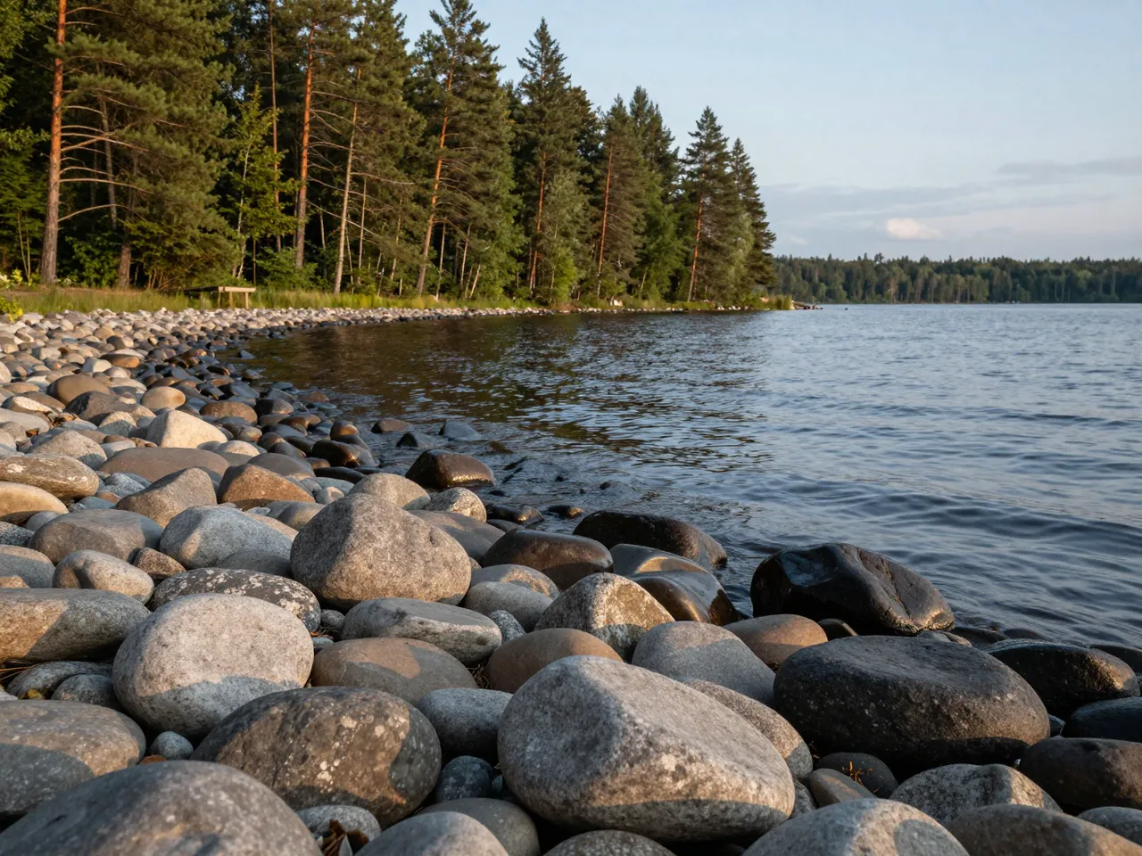 Smooth rounded stones line a calm lake shore next to a dense pine forest at late dusk