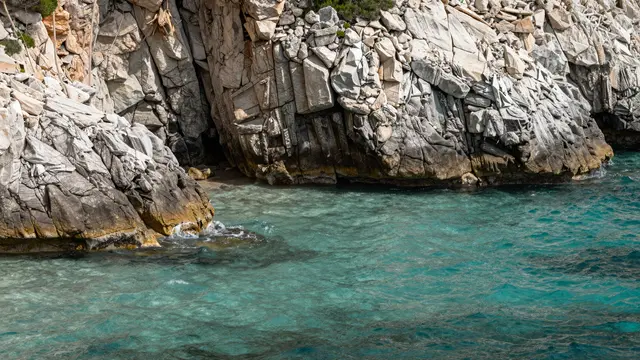 Clear turquoise water laps against rugged light gray coastal rock formations on a sunny day