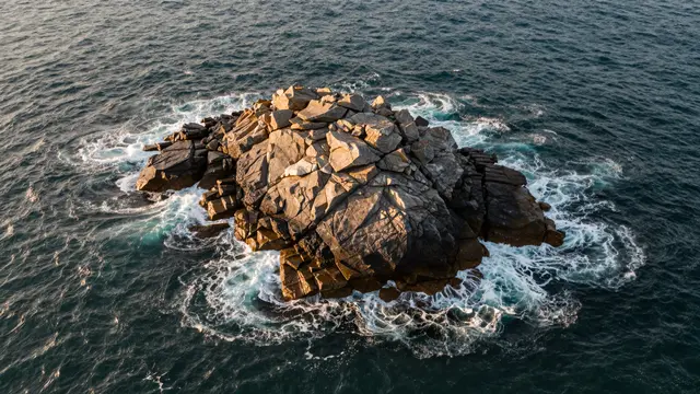 Aerial view of a rocky sea islet surrounded by foaming ocean waves at warm golden hour