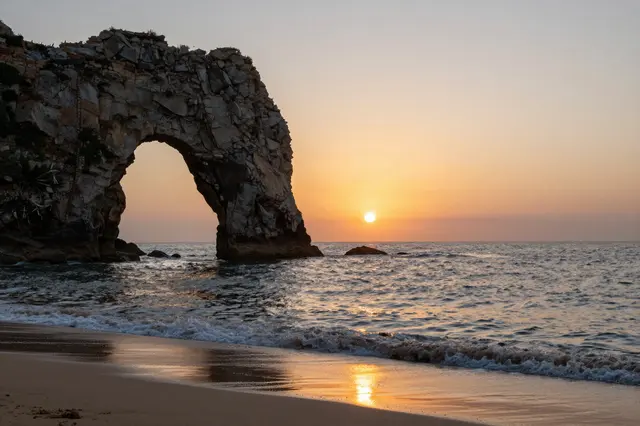 A natural rocky sea arch stands at a sandy beach as the sun sets over calm ocean waters.