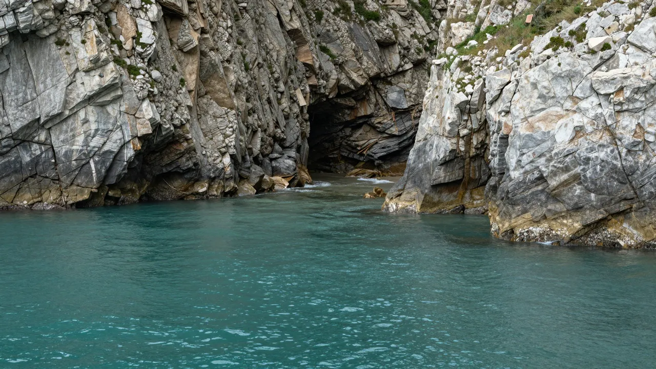 Calm turquoise water fills a narrow inlet between rugged layered gray cliffs leading to a dark sea cave.