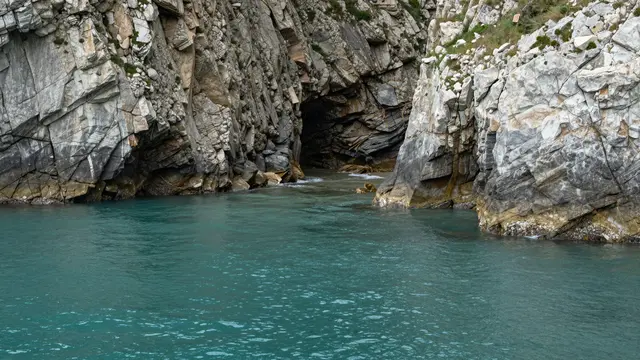 Calm turquoise water fills a narrow inlet between rugged layered gray cliffs leading to a dark sea cave.