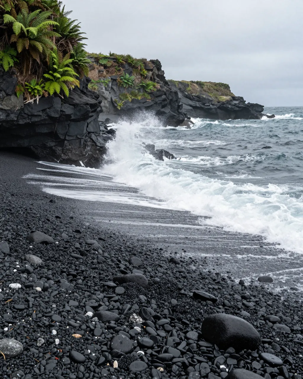 Waves crash onto a black volcanic pebble beach below fern-covered coastal cliffs under a gray overcast sky