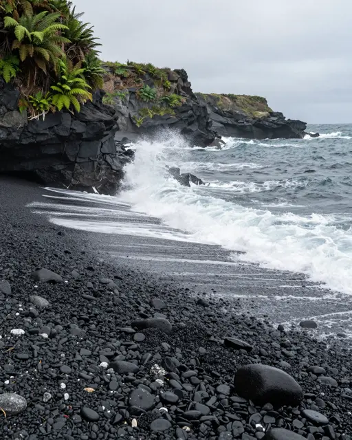 Waves crash onto a black volcanic pebble beach below fern-covered coastal cliffs under a gray overcast sky