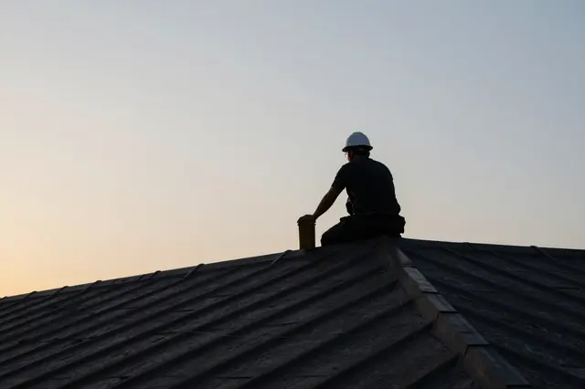 Silhouetted roofer wearing a hard hat sits on a roof ridge next to a container against a soft gradient sunrise sky