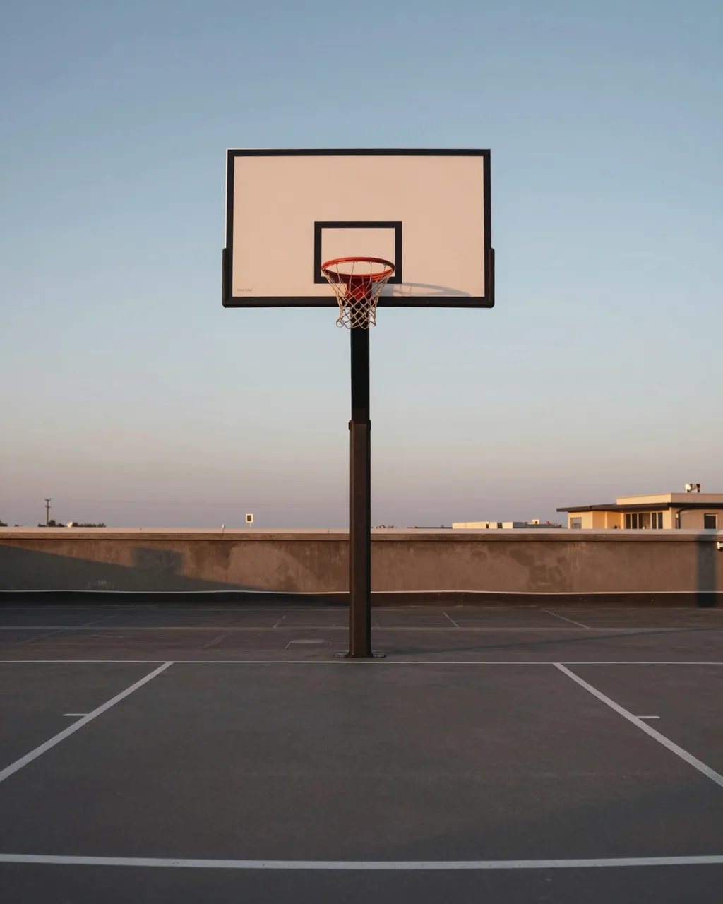 An empty rooftop basketball court with a plain backboard under a soft fading dusk sky
