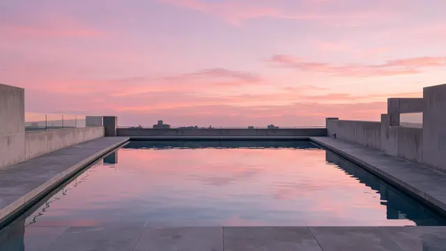 Still rooftop pool water reflects a soft pink sunset sky over concrete rooftop walls