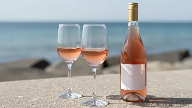 Two stemmed glasses of rosé and an unlabeled wine bottle sit on a stone ledge overlooking the calm sea